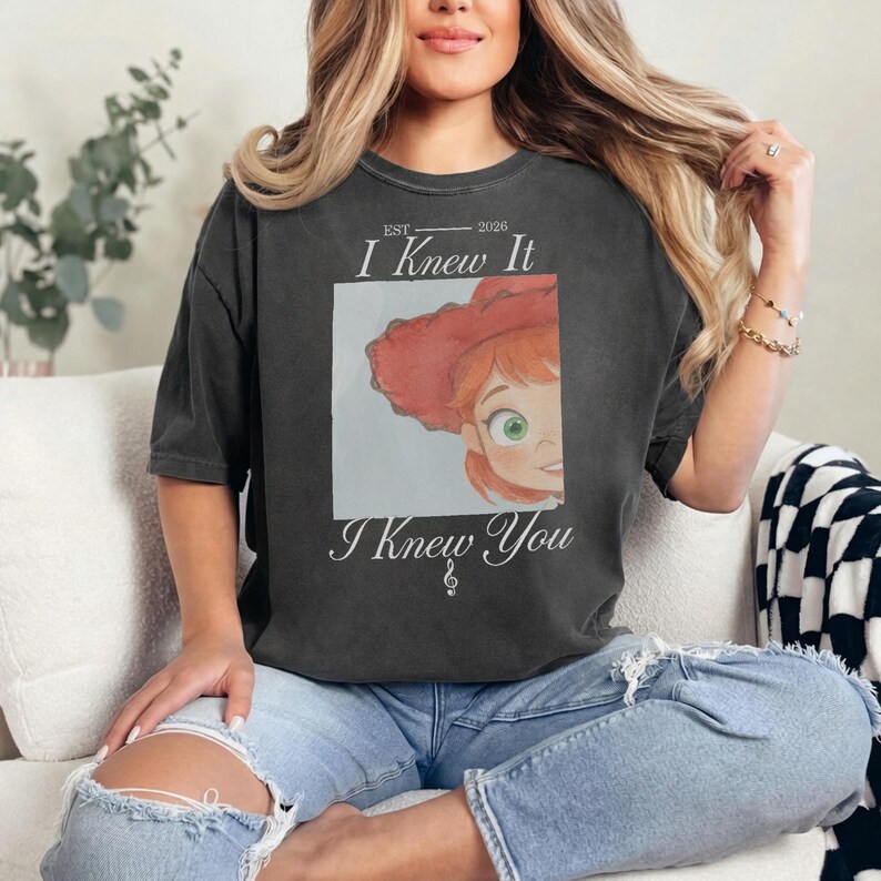 May include: A charcoal gray short-sleeve t-shirt featuring a graphic of a cartoon character with red hair and a red hat. The text reads EST 2026 I Knew It I Knew You with a small musical treble clef symbol centered below the text.