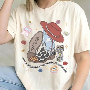 May include: A cream-colored t-shirt featuring a graphic of a cowboy boot with a cow print and blue sky pattern, a red cowboy hat, blocks with the letters T, S, and the number 13, a bow, flowers, and the text TAYLOR and I KNEW IT Y I KNEW N Y O U.