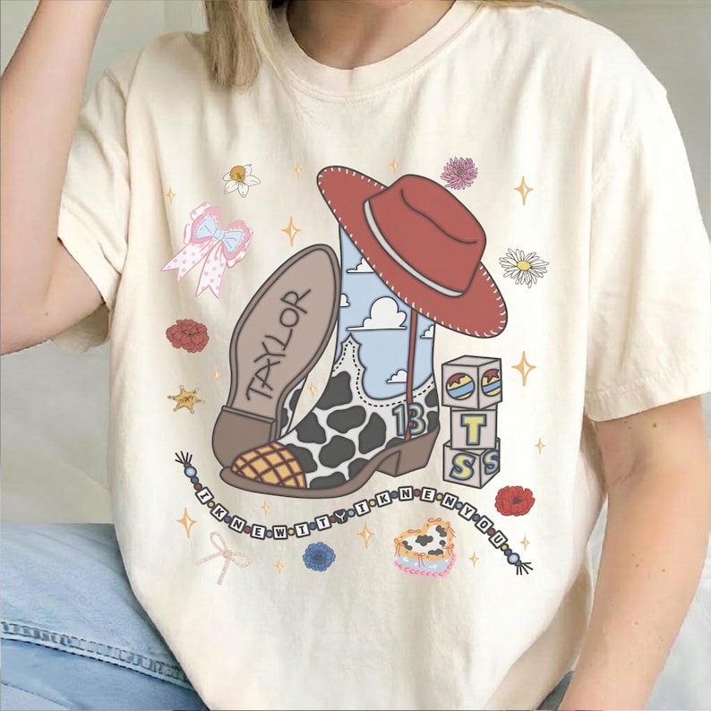 May include: A cream-colored t-shirt featuring a graphic of a cowboy boot with a cow print and blue sky pattern, a red cowboy hat, blocks with the letters T, S, and the number 13, a bow, flowers, and the text TAYLOR and I KNEW IT Y I KNEW N Y O U.