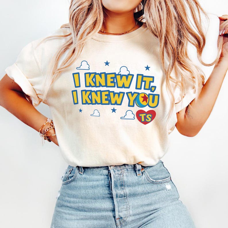 May include: A cream-colored short-sleeve t-shirt featuring the blue and yellow text I KNEW IT, I KNEW YOU with a red heart containing the letters TS, accented by blue stars and white cloud outlines, paired with light blue denim jeans.