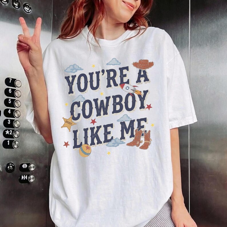 May include: A white short-sleeved t-shirt featuring the dark blue text "YOU'RE A COWBOY LIKE ME" surrounded by graphic illustrations of a brown cowboy hat, a pair of brown cowboy boots, a sheriff star badge, a rocket, clouds, and stars.