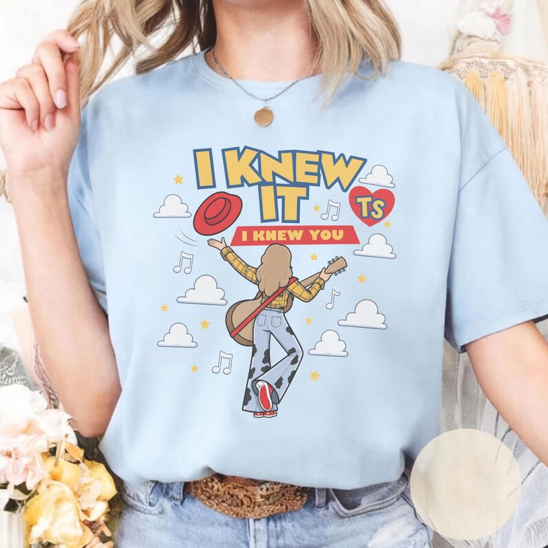 K&ouml;nnte beinhalten: Ein hellblaues T-Shirt mit einer Grafik einer Person mit langem Haar, die eine Akustikgitarre spielt und ein gelbes Karohemd sowie eine Hose mit Kuhfleckenmuster tr&auml;gt. Der Text lautet I KNEW IT I KNEW YOU mit einem roten Hut und Wolken.