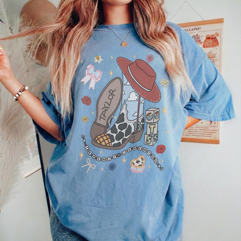 May include: A blue t-shirt featuring a graphic print of a cow-print cowboy boot with the name Taylor, a red cowboy hat, blocks with the letters T S, a bow, flowers, and a beaded string with text. The design includes stars and clouds.