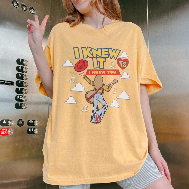 K&ouml;nnte beinhalten: Ein gelbes T-Shirt mit einer Grafik einer Person mit langen Haaren, die Gitarre spielt und ein kariertes Hemd sowie eine Hose mit Kuhfleckenmuster tr&auml;gt. Das Design umfasst einen roten Hut, wei&szlig;e Wolken, Noten, ein rotes Herz mit TS und den Text I KNEW IT I KNEW YOU.