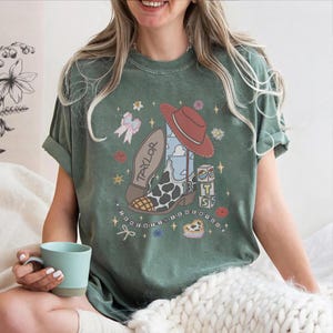 May include: A sage green t-shirt featuring a graphic print of a cowboy boot with a cow print pattern, a red hat, a bow, and blocks with the text "T S". The design includes the name "TAYLOR" on the boot sole and the text "I KNEW YOU WERE TROUBLE".