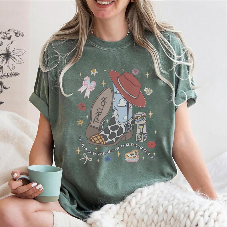 May include: A sage green t-shirt featuring a graphic print of a cowboy boot with a cow print pattern, a red hat, a bow, and blocks with the text "T S". The design includes the name "TAYLOR" on the boot sole and the text "I KNEW YOU WERE TROUBLE".