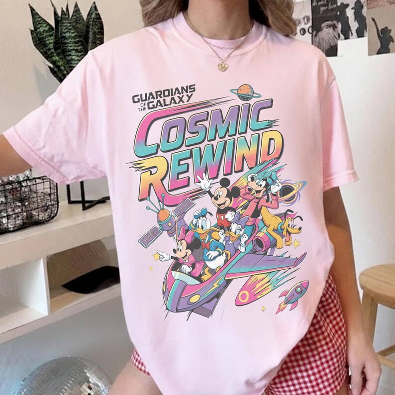 Puede incluir: Una camiseta de manga corta de color rosa claro con un estampado gr&aacute;fico que incluye el texto Guardians of the Galaxy Cosmic Rewind. El dise&ntilde;o presenta ilustraciones coloridas de Mickey Mouse, Minnie Mouse, Donald Duck, Daisy Duck, Goofy y Pluto.