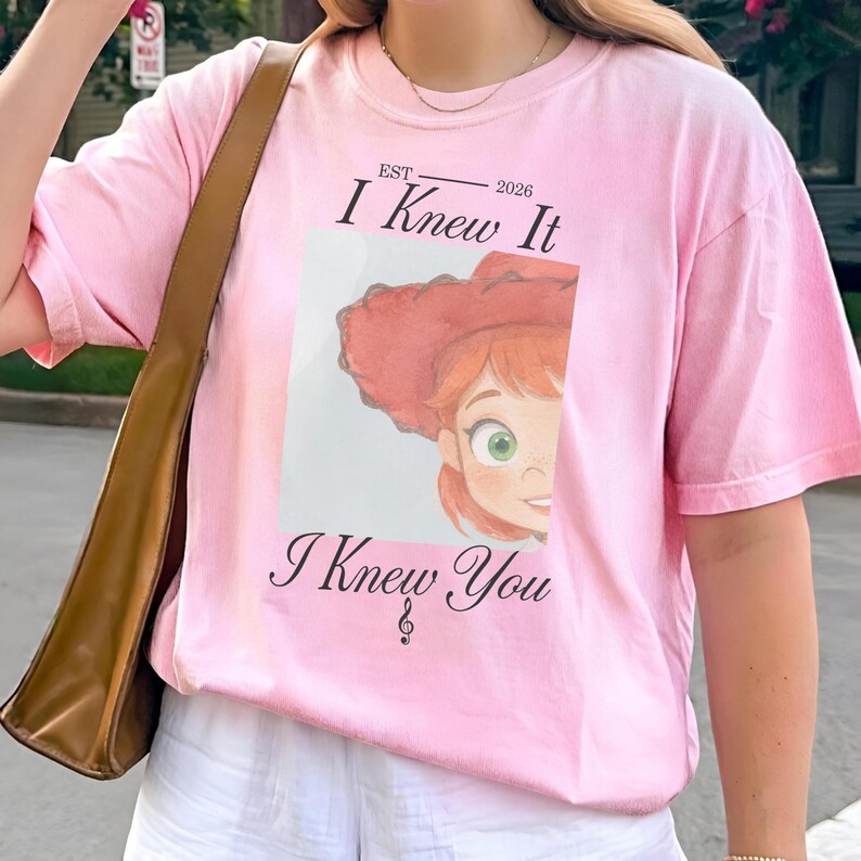 May include: A light pink short-sleeve t-shirt featuring a graphic of a cartoon character with red hair and a red hat. The text reads "EST 2026 I Knew It" above the graphic and "I Knew You" below it, with a small treble clef symbol included.