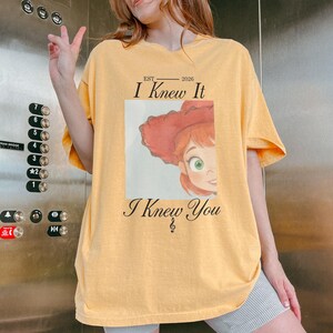 May include: A light yellow short-sleeve t-shirt featuring a graphic of a red-haired character wearing a red hat. The text reads EST 2026 I Knew It I Knew You with a treble clef symbol below the text. The design is centered on the chest area.