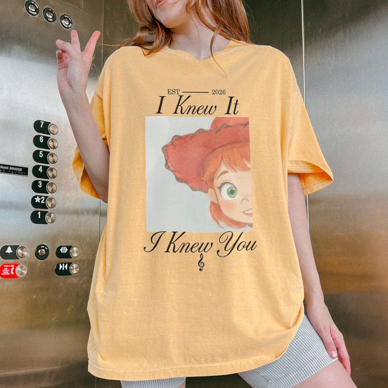 May include: A light yellow short-sleeve t-shirt featuring a graphic of a red-haired character wearing a red hat. The text reads EST 2026 I Knew It I Knew You with a treble clef symbol below the text. The design is centered on the chest area.