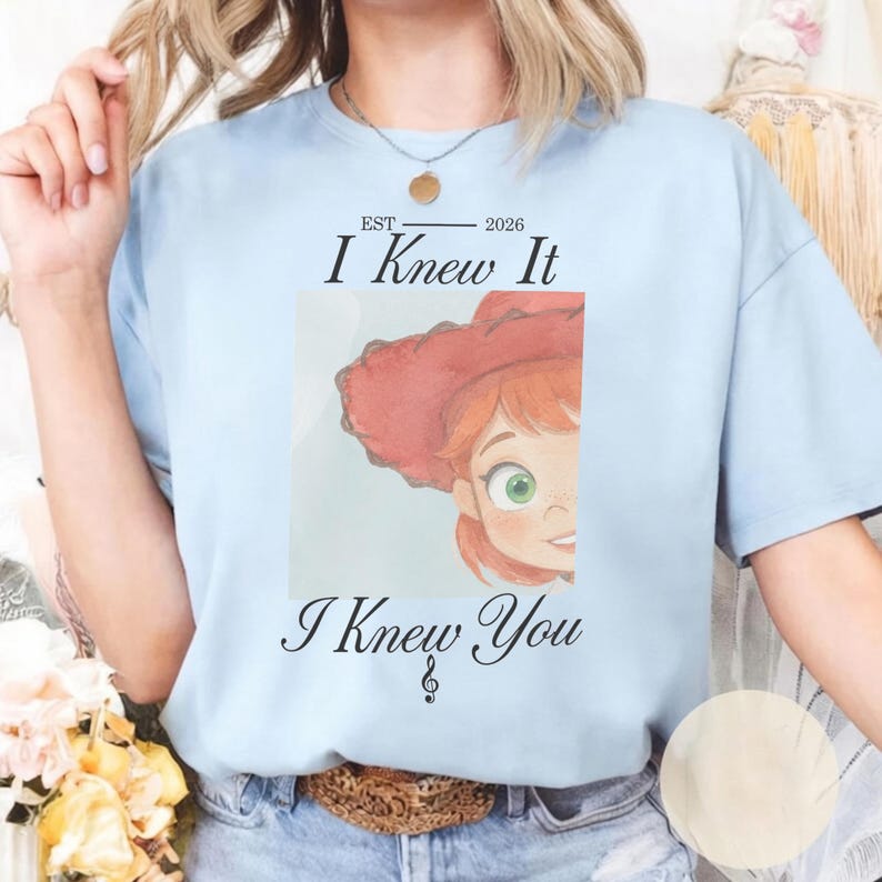 May include: A light blue t-shirt featuring a graphic of a cartoon character with red hair and a red hat. The text reads EST 2026 I Knew It I Knew You with a treble clef symbol below the text. The design is centered on the front of the shirt.