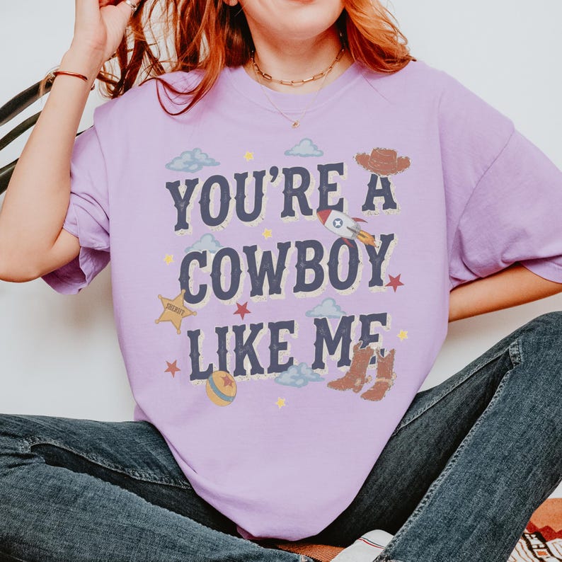 May include: A light purple short-sleeve t-shirt featuring the dark blue text "YOU'RE A COWBOY LIKE ME" surrounded by graphic illustrations of a rocket, a sheriff star, a cowboy hat, cowboy boots, a ball, clouds, and small yellow stars.
