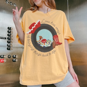 May include: A yellow short-sleeve t-shirt featuring a graphic of a black tire framing cartoon characters, a red cowboy hat, and a red boot. The text reads, "Love has ways of bringing things back to life" and "TAYLOR" is printed on the red boot.