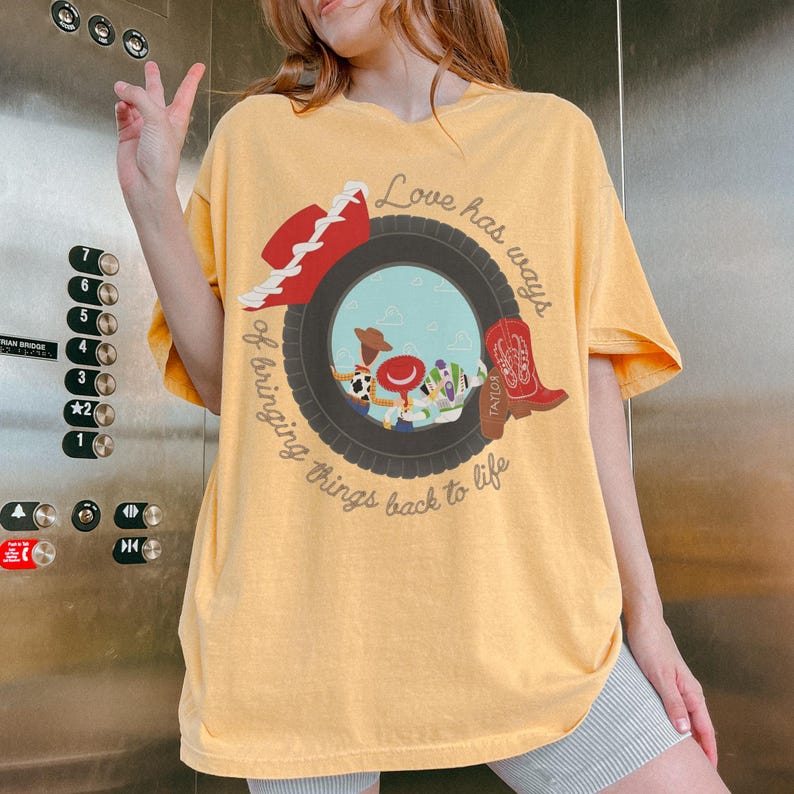 May include: A yellow short-sleeve t-shirt featuring a graphic of a black tire framing cartoon characters, a red cowboy hat, and a red boot. The text reads, "Love has ways of bringing things back to life" and "TAYLOR" is printed on the red boot.