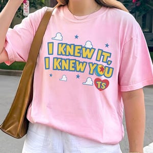 May include: A light pink short-sleeve t-shirt featuring the yellow and blue text I KNEW IT, I KNEW YOU with a red heart containing the letters TS, surrounded by small blue stars and white cloud graphics printed on the front of the garment.
