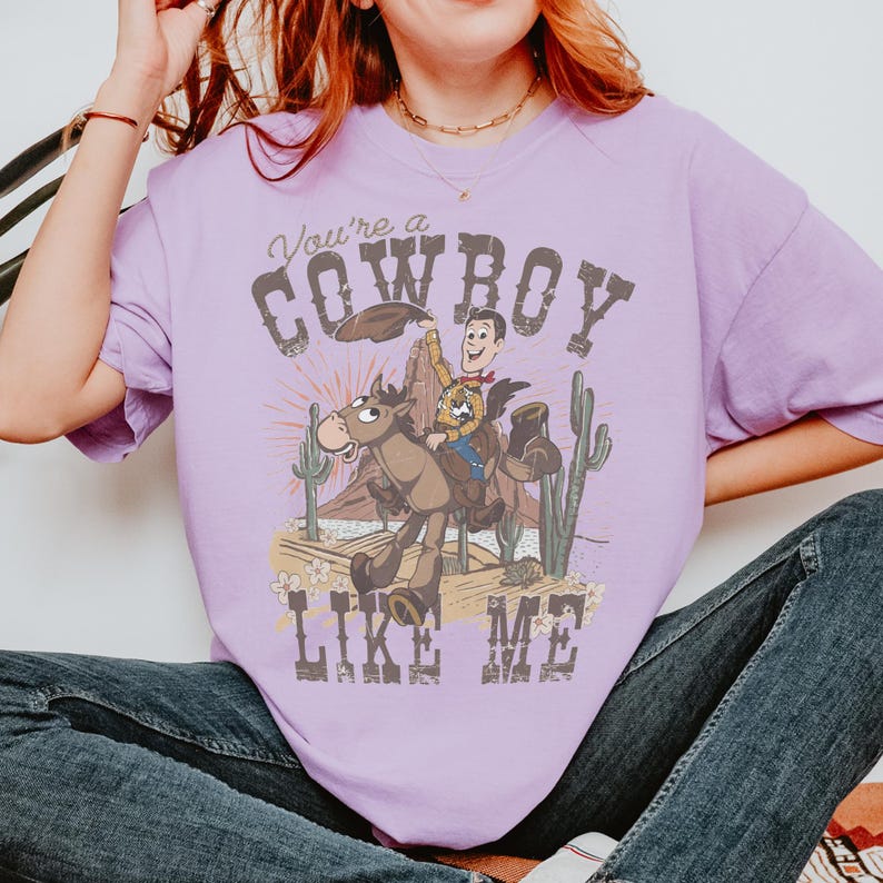 Puede incluir: Una camiseta de manga corta de color morado claro con un estampado gr&aacute;fico de un vaquero de dibujos animados montando a caballo en un paisaje des&eacute;rtico con cactus y monta&ntilde;as. El texto "You're a COWBOY LIKE ME" est&aacute; impreso en una fuente desgastada.
