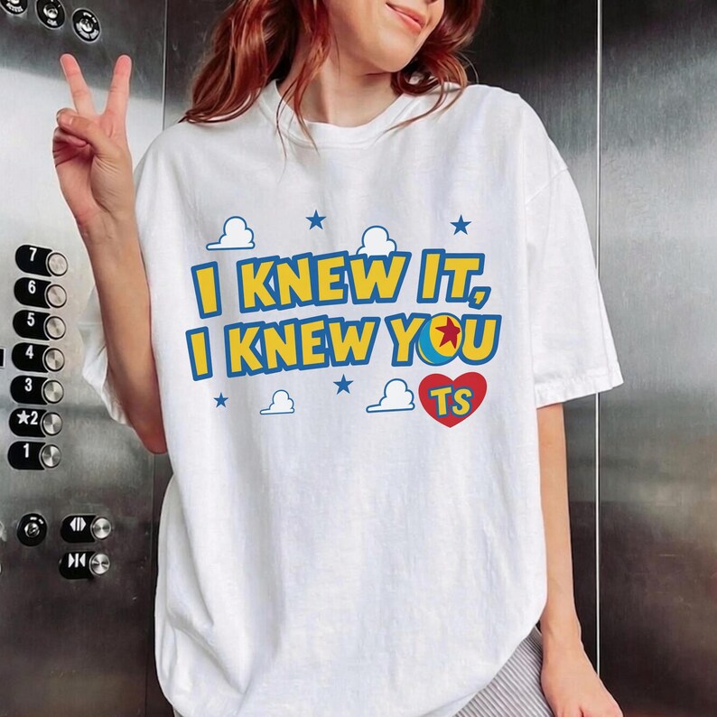 May include: A white short-sleeved t-shirt featuring the blue and yellow text I KNEW IT, I KNEW YOU, a red heart with the letters TS, and a graphic of a ball with a red star, surrounded by small blue stars and white cloud outlines on the front.