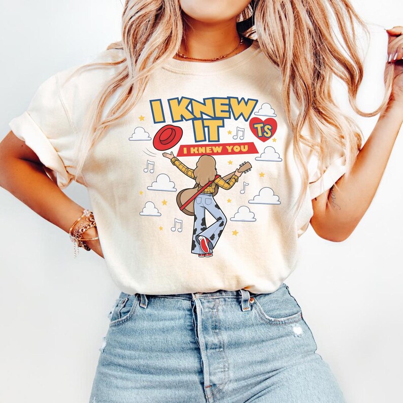 K&ouml;nnte beinhalten: Ein cremefarbenes T-Shirt mit einer grafischen Illustration einer Person, die eine Akustikgitarre spielt. Der Text lautet I KNEW IT und I KNEW YOU, begleitet von einem roten Hut, einem roten Herz mit TS, Musiknoten, Sternen und wei&szlig;en Wolken.