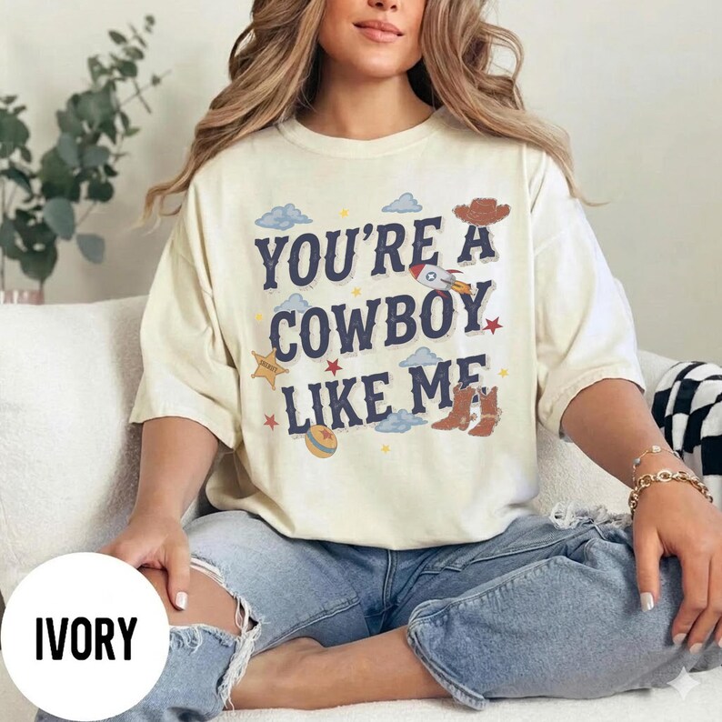 May include: An ivory short-sleeve t-shirt featuring the dark blue text "YOU'RE A COWBOY LIKE ME" surrounded by graphic illustrations of a brown cowboy hat, a rocket ship, a sheriff star badge, a colorful ball, and brown cowboy boots.