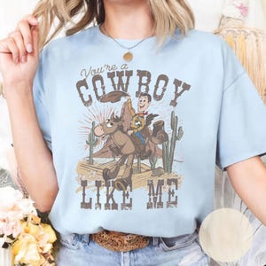 Puede incluir: Una camiseta azul claro con un estampado gr&aacute;fico de un vaquero de dibujos animados montando a caballo en un paisaje des&eacute;rtico con cactus y formaciones rocosas. El texto "You're a COWBOY LIKE ME" aparece en una fuente marr&oacute;n desgastada.