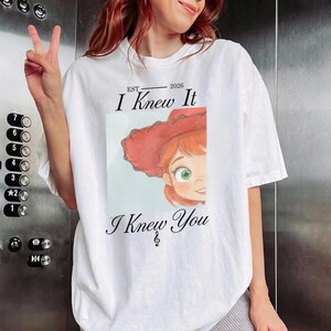 May include: A white short-sleeved t-shirt featuring a graphic of a cartoon character with red hair and a red hat. The text reads EST 2026 I Knew It I Knew You with a small musical note symbol below the text. The design is centered on the chest.
