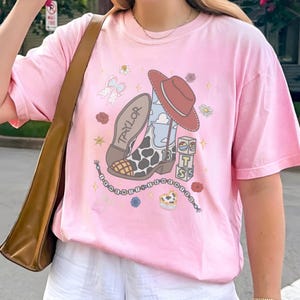 May include: A light pink t-shirt featuring a graphic print of a cowboy boot with a cow print pattern, a red hat, a stack of blocks with the letters T, S, and a sun, and a beaded string. The boot has the name TAYLOR and the number 13 printed on it.