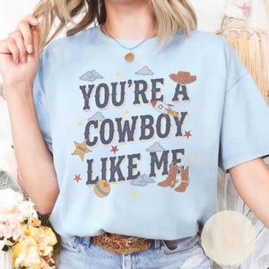 May include: A light blue short-sleeve t-shirt featuring the dark blue text "YOU'RE A COWBOY LIKE ME" surrounded by graphic illustrations of a rocket, a sheriff star, cowboy boots, a ball, clouds, and small stars in various colors.