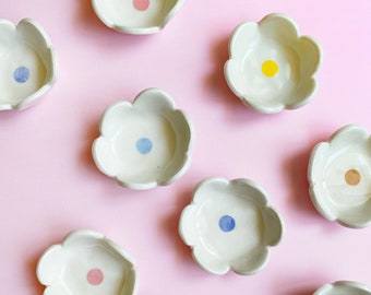 May include: A collection of small, off-white ceramic dishes shaped like five-petaled flowers. Each dish features a single, centered circular dot in various colors, including pink, yellow, light blue, and tan, arranged in a scattered pattern.
