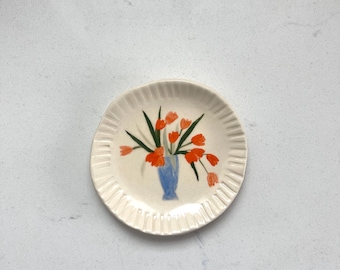 May include: A round, off-white ceramic plate with a fluted, scalloped edge. The center of the plate features a hand-painted illustration of a blue vase filled with a bouquet of vibrant orange tulips and green stems and leaves.