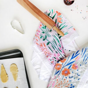 May include: A baking sheet with a white parchment paper liner and cut-out cookie dough in the shape of a bird. A rolling pin, a cookie cutter, and a bowl of sprinkles are also visible.