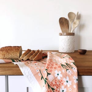 May include: A pink tea towel with a floral pattern in shades of green, white, and orange. The towel is folded and draped over a wooden countertop with a loaf of sliced bread on top.