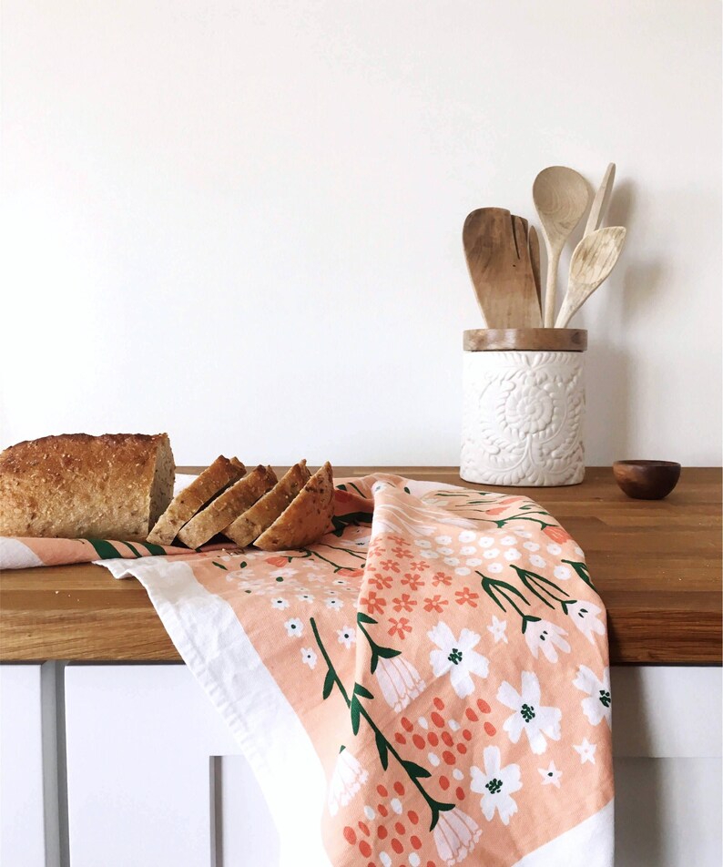 May include: A pink tea towel with a floral pattern in shades of green, white, and orange. The towel is folded and draped over a wooden countertop with a loaf of sliced bread on top.