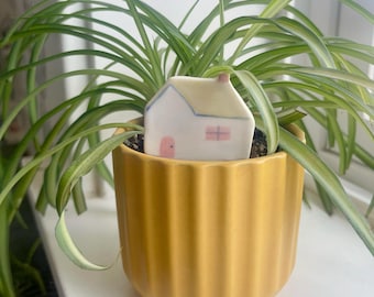 May include: A small ceramic house ornament with a white finish, a pink door, and a pink window, featuring blue roof lines. The house is placed inside a vibrant yellow, vertically ribbed cylindrical plant pot containing a green spider plant.