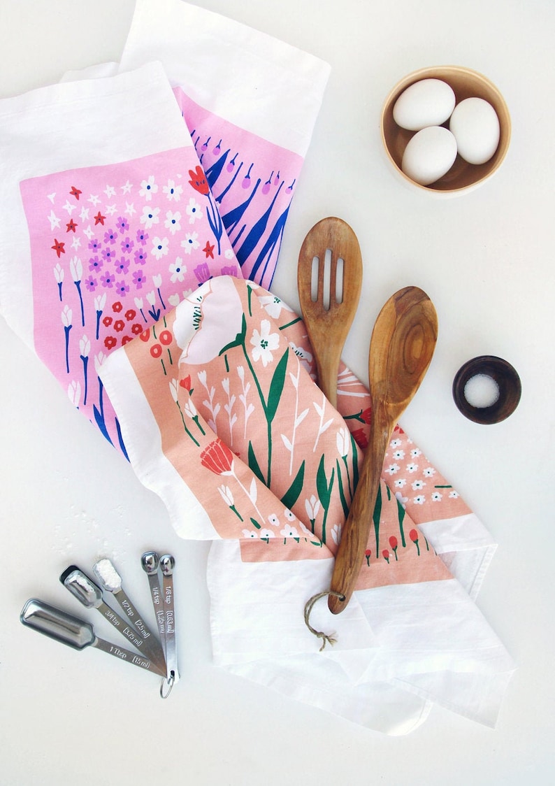 May include: Two pink and white floral tea towels with a white border, a wooden spoon, a slotted wooden spoon, a small bowl with three white eggs, and a set of four measuring spoons.