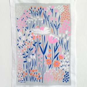 May include: A gray tea towel with a floral pattern in shades of pink, orange, and blue. The pattern features a variety of flowers and leaves, including daisies, tulips, and ferns. The text "Jean Cooper" is printed in the bottom left corner of the towel.