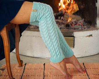 May include: Light blue cable knit leg warmers with a small wooden tag. The leg warmers are worn over the knees. The background includes a fireplace with a burning fire and a wooden chair.
