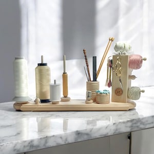 May include: A wooden sewing organizer with various spools of thread, needles, scissors, and other craft supplies. The organizer includes a spool holder, a pin cushion, and small wooden containers. The items are arranged on a light-colored wooden tray.