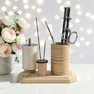 May include: A light-colored wooden sewing organizer base holding a large spool of white thread, a small wooden spool with two metal needles, and a cylindrical wooden cup containing a metal ruler and silver scissors for craft storage.