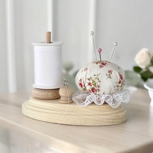 May include: A wooden sewing station with a spool of white thread, a floral pin cushion with silver pins, and a small wooden thread holder. The pin cushion has a white lace trim and is decorated with red and green floral patterns.