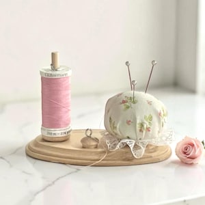 May include: A wooden sewing station with a spool of pink thread, a floral pin cushion with pins, and a small wooden thread holder. The thread spool reads "Gütermann". A pink rose is next to the station.