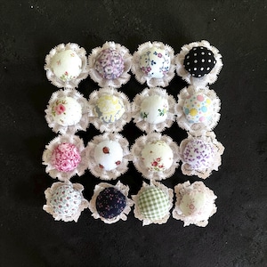 May include: A collection of sixteen fabric-covered buttons arranged in a four-by-four grid. Each button features a unique pattern, including floral prints, polka dots, gingham, and ladybugs, all framed by a delicate white lace trim border.