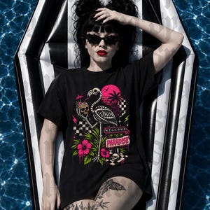 May include: Black t-shirt with a graphic of a flamingo skeleton, skull, and the words "Welcome to Paradise." The shirt is worn on a black and white striped pool float in a pool. The person is wearing sunglasses and has tattoos.