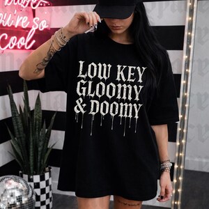 Puede incluir: Camiseta negra de gran tamaño con el texto "LOW KEY GLOOMY & DOOMY" en letras góticas blancas. La camiseta se combina con botas negras y calcetines a cuadros. Un letrero de neón dice "babe you're so cool".
