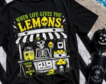 May include: A black t-shirt featuring a graphic of a skeleton running a lemonade stand. The text reads "WHEN LIFE GIVES YOU LEMONS, THROW THEM AT PEOPLE" and "$1 PER LEMON". The design includes yellow lemons, a jar, a bucket with a skull, and a striped awning.