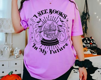 May include: A lilac-colored t-shirt with the text "I See Books In My Future" printed on the front. The graphic features a crystal ball with books inside, held by hands with long nails. The shirt is a casual, short-sleeved style.