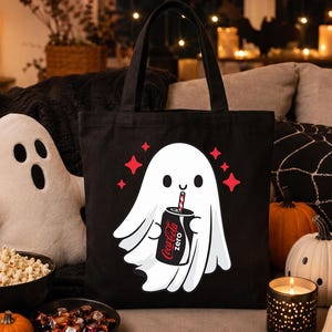 May include: A black tote bag featuring a white cartoon ghost illustration holding a black Coca-Cola Zero can with a red and white striped straw. The ghost is surrounded by small red four-pointed star shapes on the front of the fabric bag.