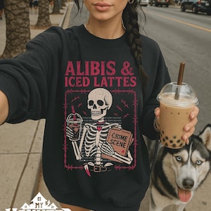 May include: Black sweatshirt with the text "ALIBIS & ICED LATTES" and a skeleton graphic holding a drink and a book labeled "CRIME SCENE". A husky dog with blue eyes is next to the person wearing the sweatshirt.