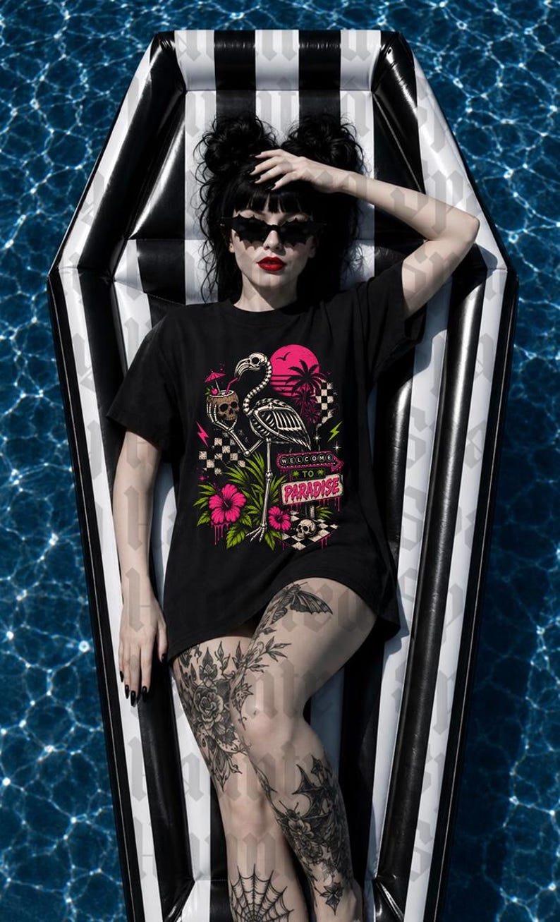 May include: Black t-shirt with a graphic of a flamingo skeleton, skull, and the words "Welcome to Paradise." The shirt is worn on a black and white striped pool float in a pool. The person is wearing sunglasses and has tattoos.