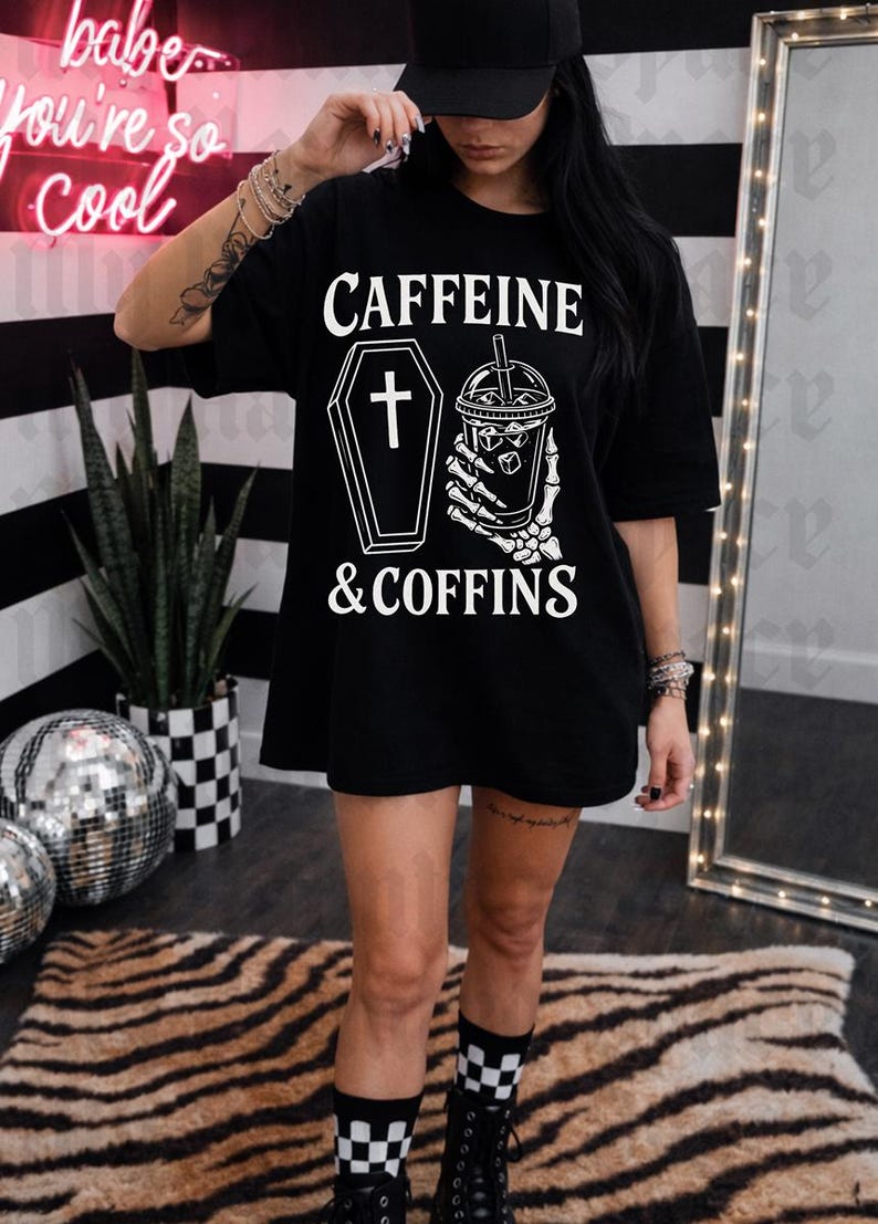 May include: Black t-shirt with the text "CAFFEINE & COFFINS" in white. The design includes a coffin with a cross and a skeleton hand holding a coffee cup. The person is wearing a black cap and black boots.