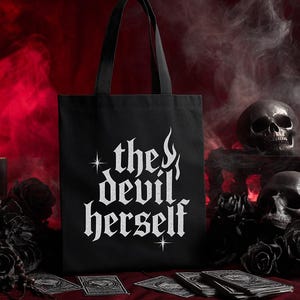 Puede incluir: Bolso tote negro con el texto blanco "the devil herself" en letras góticas. El bolso tiene asas negras y se exhibe sobre una superficie roja con calaveras y rosas negras en el fondo. Un montón de cartas del tarot está frente al bolso.